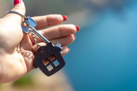 Keys with a keychain in the shape of a house in a female hand against the backdrop of the sea. The concept of buying a house, apartment, real estate, young family, mortgage loan, rental housing