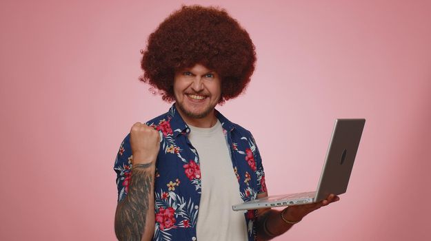 Excited happy tourist man typing on laptop computer, working on project, enjoying results, winning lottery, celebrating success, online shopping. Young bearded guy boy isolated on blue background