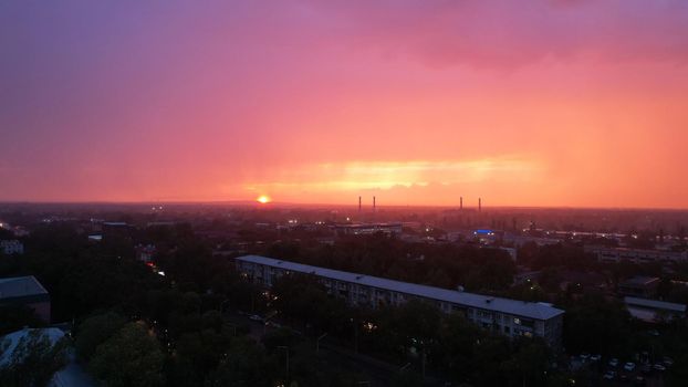 Epic sunset with clouds over the city of Almaty. The gradient of clouds is from dark blue to purple-orange. Green tall trees, houses, a road with cars. The lights are on. It's raining heavily