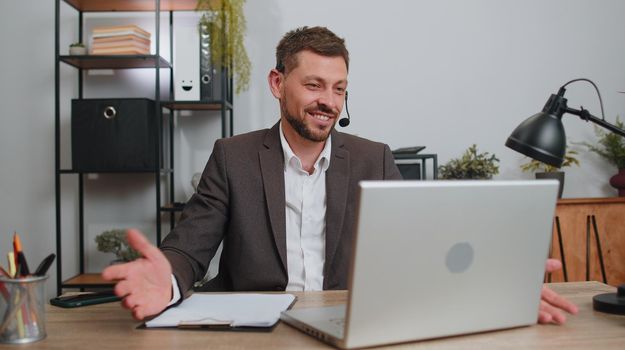 Businessman working on laptop computer wearing headset, freelance worker call center or support service operator helpline, having pleasant talk with client or colleague communication support at office