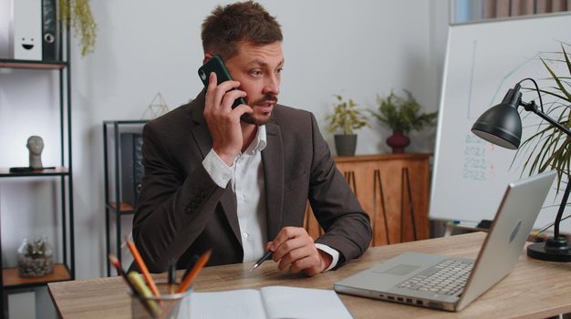 Conflict, quarrel, negotiation. Angry businessman ceo in formal wear suit making phone call at home office desk. Concentrated upset mad male freelancer man talking smartphone conversation indoors