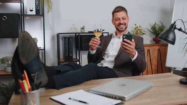 Cheers. Businessman drinking champagne from glass after working, putting legs on table sitting at office. Happy male manager freelance man celebrate successful contract agreement holding smartphone
