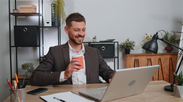 Businessman freelancer enters office start working on laptop computer at desk and drinking morning coffee. Man works on notebook, sends messages makes online purchases watching social media movies