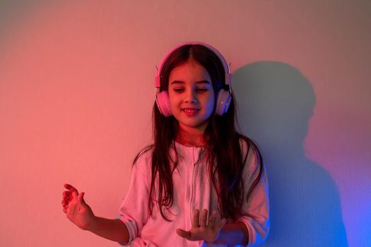 Little girl in pink headphones and cozy clothes dances to music in neon pink blue light