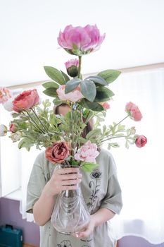 Teenager girl holding a bunch of pink and red peonies in glass vase while closing her face with branches. High quality photo