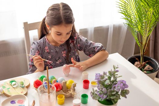 A adorable little girl sits at a white table near the window, prepares for the Easter holiday, paints eggs with a finger.