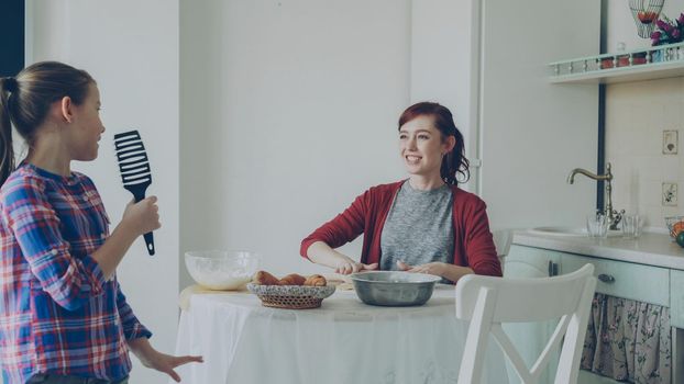 Young mother rolling dough while cooking and her little funny daughter walking around and singing with comb dancing in the kitchen on weekend. Family, food, home and people concept