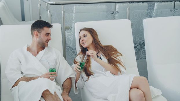 Young lovers in bathrobes are relaxing in chairs in spa salon, drinking cocktails and talking. Wellness, romantic relationship and happiness concept.