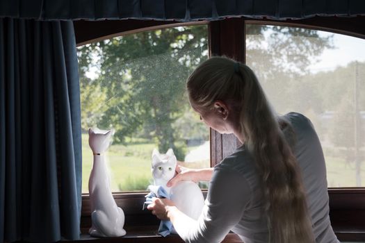 a woman wipes a porcelain figurine of a white cat on the windowsill,room cleaning and tidying up. High quality photo