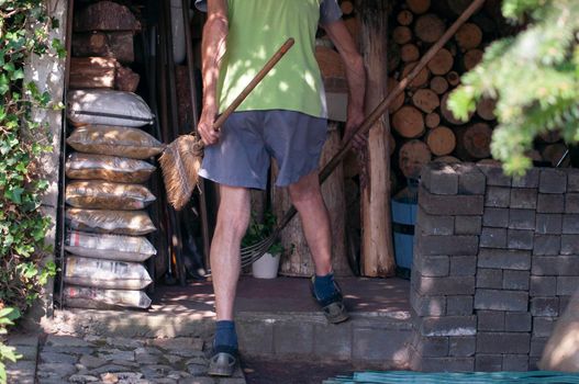 gardener in dirty work clothes and boots holds garden tools, plastic brush broom, seasonal spring work in the garden. High quality photo