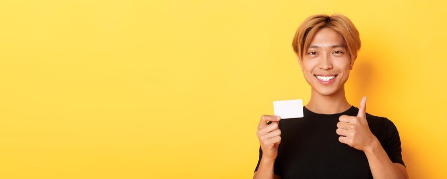 Close-up of satisfied asian guy recommend bank, showing credit card and standing over yellow background.
