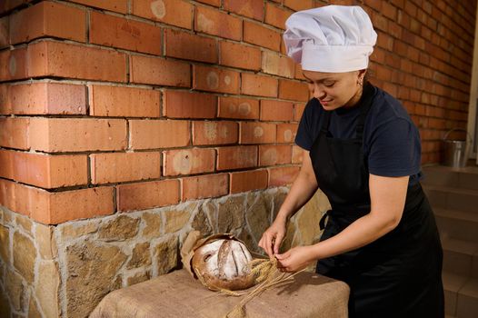Female baker chef in a black apron and white cap, places wheat spikelets next to freshly baked whole grain bread on a table covered with burlap tablecloth, putting it up for sale with fresh pastries