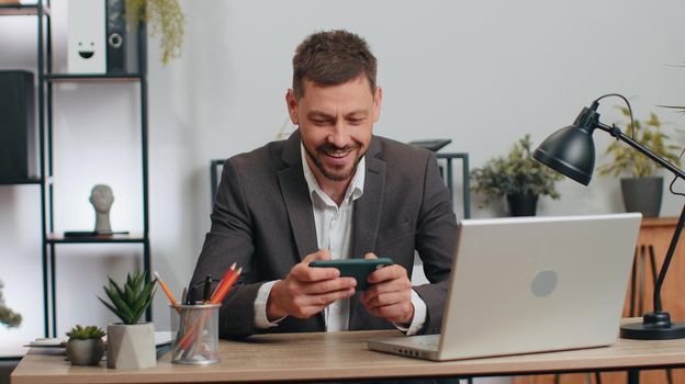 Businessman working with documents at office desk, taking a break, playing smartphone game. Male manager freelancer man winner celebrate mobile phone online win victory success receive read good news