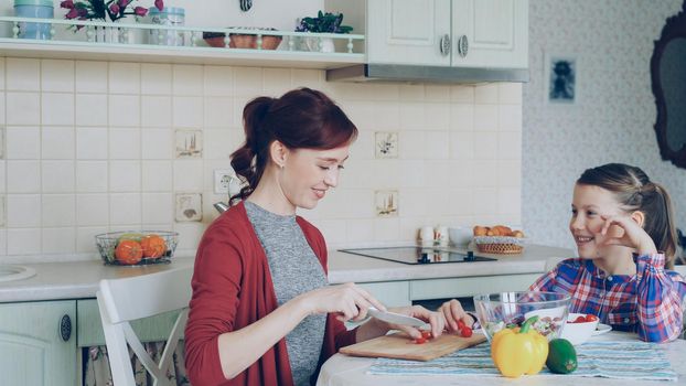 Cheerful happy mother cutting tomatoes while cooking and talking to her smiling daughter feeding mom in the kitchen at home. Family, cook, and people concept