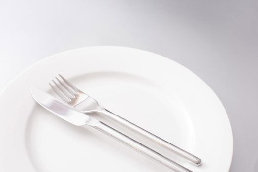 Clean generic white dinner plate with silver knife and fork neatly arranged in the centre in a close up cropped view