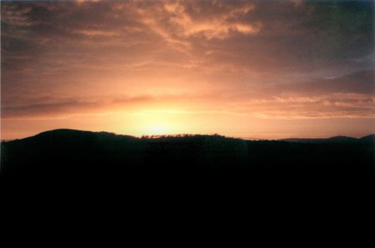 Fiery orange sunset over silhouetted mountains as the sun sinks below the horizon with the glow lighting up the clouds