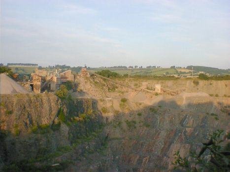 Artificial landscape of a big old quarry with green plants growing on the steep slopes and factory facilities on top, viewed from across