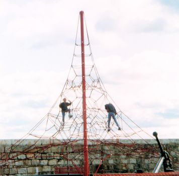 Two young men climbing on a exterior tall play frame in front of an old stone wall an overcast sky