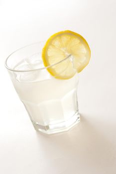 Iced drink in a glass tumbler garnished with a slice of tangy lemon on white, clear liquid at a tilted angle