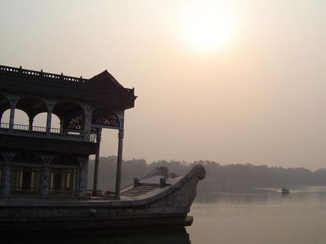 Detail of House Boat with Ornate Carvings Floating on Calm River with Small Boat and Shoreline in Background Under Hazy Sky with Obscured Sun
