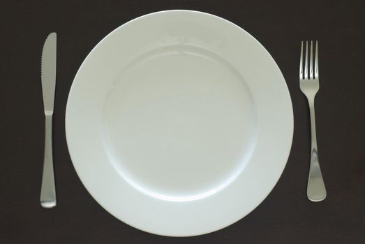 Plain white clean empty dinner plate and cutlery place setting on a black table viewed from above