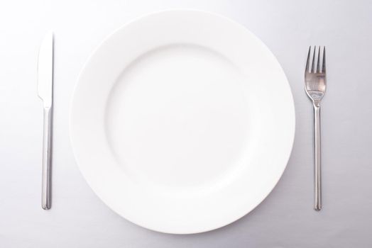 Clean empty white dinner plate with silver metal knife and fork in a simple place setting ready to serve food