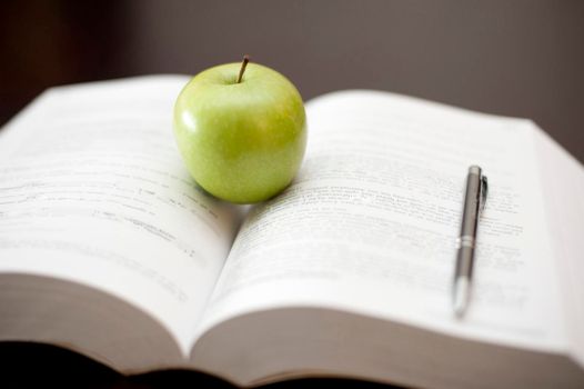 A fresh juicy green apple stands on an open textbook with a pen conceptual of education and learning