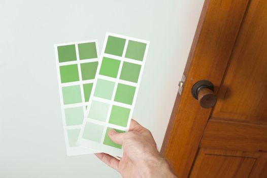 Man holding up cards of green paint swatches against a white wall in his house as he makes a decision on colors to redecorate the interior