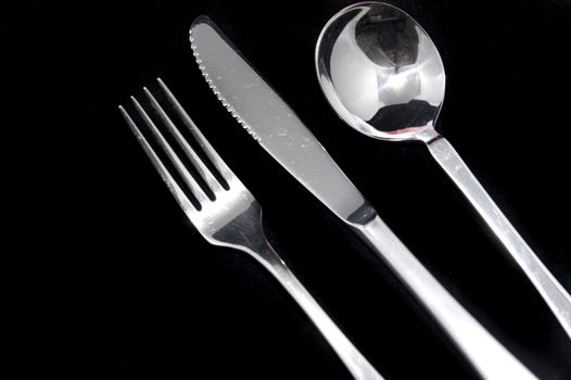 Clean metal knife, fork and spoon on black background with copy space arranged diagonally from the left corner