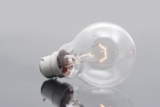 Domestic light bulb with a bayonet fitting and a glowing filament on a reflective grey surface for power and energy or bright ideas concepts