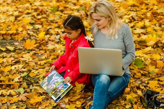 Happy smiling mother and child sits together looking on book or tablet pc in autumn park