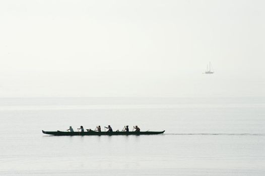 Six men team training for a race in their canoe paddling in unison on a calm ocean, silhouette against a misty sea