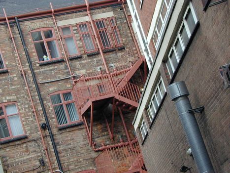 Red metal fire escape on the exterior of a an old face brick building viewed at a tilted angle from below