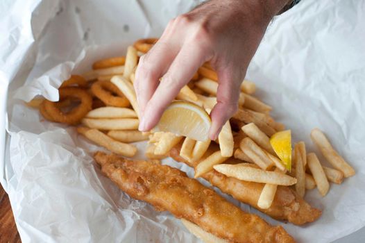 Hand squeezing lemon over takeaway fried fish fillets and calamari in batter with fried potato chips on crumpled paper