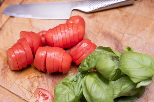 Sliced red raw tomatoes and fresh basil leaves in close-up on chopping board with kitchen knife in background