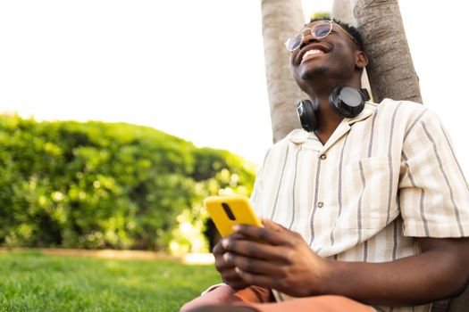 Happy black man relaxing outdoors using mobile phone. College student laughing on campus. Copy space. Lifestyle concept.