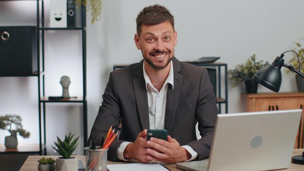 Business man programmer software developer in suit working at office finishing up phone call selling online using smartphone making victory gesture, receiving good news rejoicing over success triumph