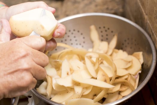 Man peeling fresh potatoes for cooking catching the peels in a metal colander, close up on his hands and the utensil