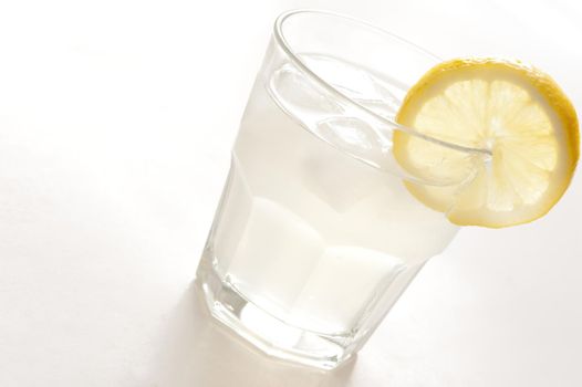 Long glass of clear spirits served with ice and garnished with a slice of fresh lemon, tilted angle close up on white