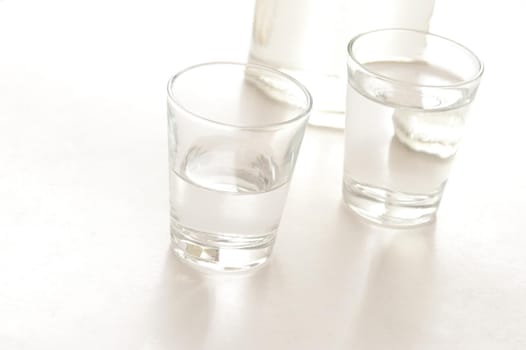 Two shot glasses with servings of white spirits, possibly neat vodka, with the base of a bottle of alcohol in the background