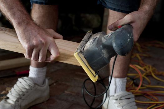 Handyman doing joinery using an electric sander to smooth the surface of a plank of wood before installing it in a house, close up of his hands