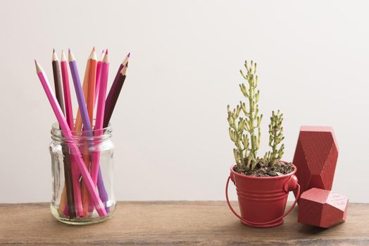 Desk still life with red ornaments and a potted plant in a little bucket alongside a jar of colored pencils against a white wall background