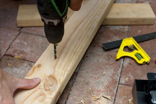 Carpenter drilling a hole in a plank of wood using a handheld electric power drill as he works on a brick floor
