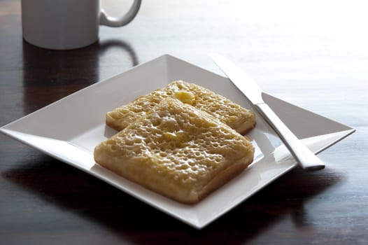 Modern breakfast of two square crumpets with knife on a white plate besides a coffee mug