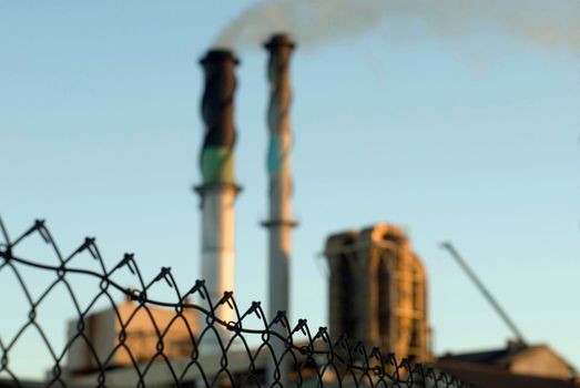 Industrial background of a smoking chimney at an industrial manufacturing plant causing air pollution protected by a wire mesh perimeter fence with focus to the fence