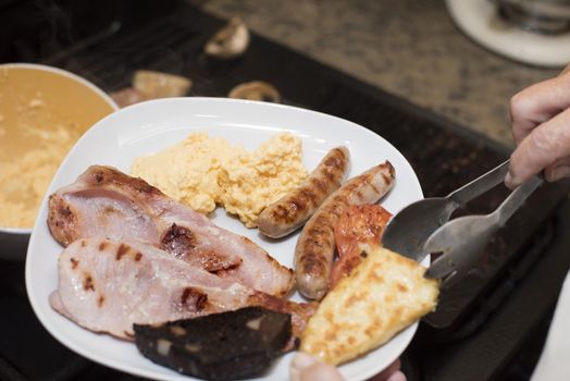 Hand using stainless steel tongs to place a hash brown beside sausage and ham on breakfast plate