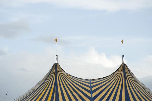Striped Big Top circus tent with colorful alternating yellow and blue stripes and flags on the poles against a cloudy sky