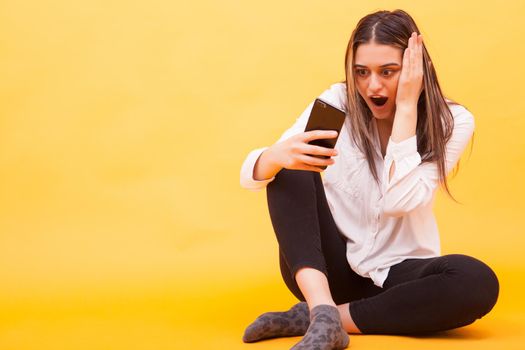 Girl looking amazed at her phone while sitting down over yellow background. Facial expression