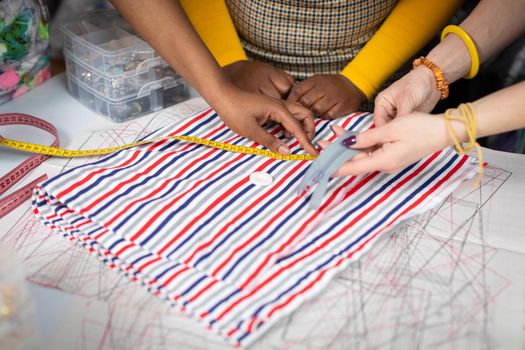 The striped fabric is lying on the tailor's table and the seamstress is measuring whether she has enough to sew the dress. Two African women and a European woman in a tailor's shop.