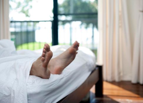 Bare male feet protruding from under the white bedclothes at the end of a bed as a man enjoys a relaxing lie in on a sunny day